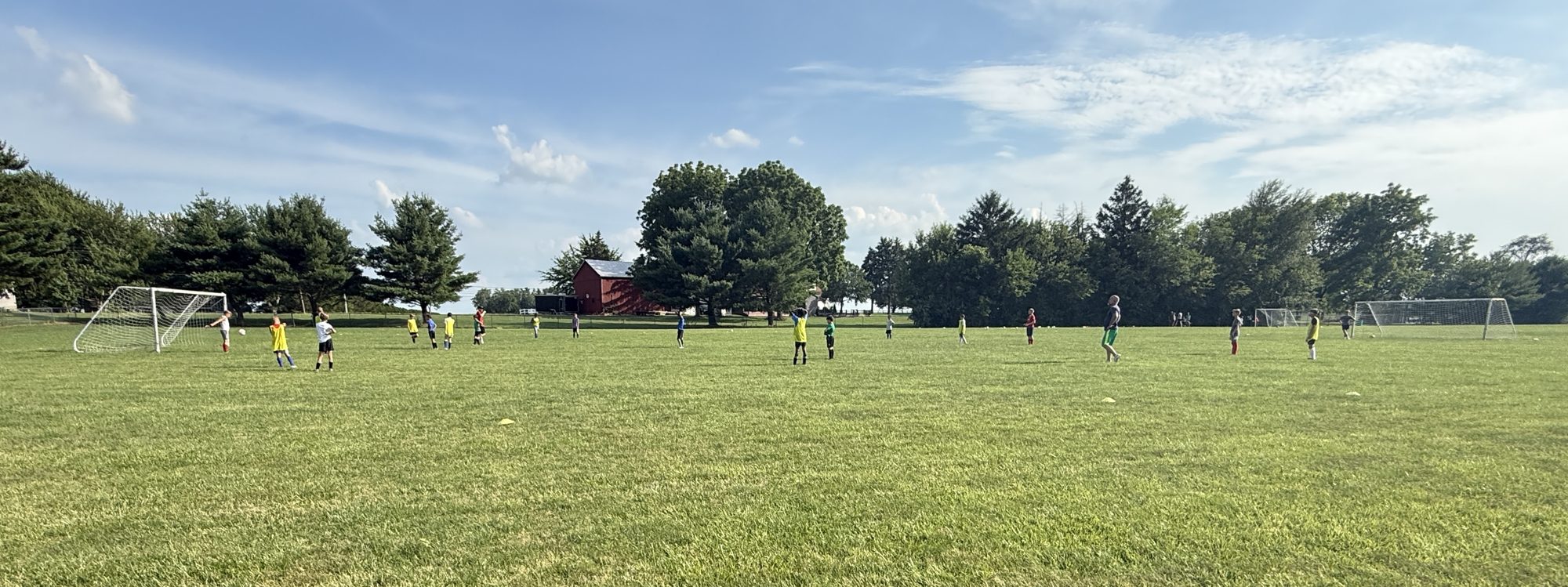 Kids of many ages in yellow, blue, red and green pinnies spread across the Liberty-Benton grass field at a session, with the district's red barn and tree line behind.